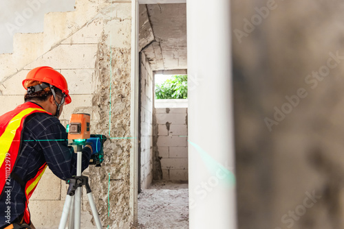 Construction Worker with Laser Level A skilled construction worker, equipped with a hard hat and safety vest, precisely uses a laser level to ensure accurate measurements on a building site.