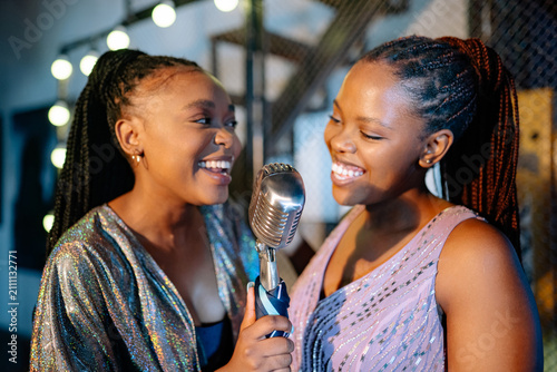 Canvas Print Two cheerful young Black women performing with a retro microphone during an exciting evening event in the industrial jazz bar setting