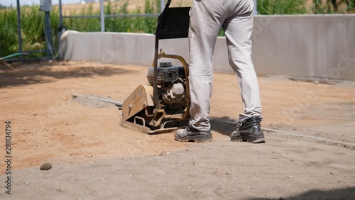 Fotografie Worker in work pants and safety boots uses plate compactor to flatten soil near