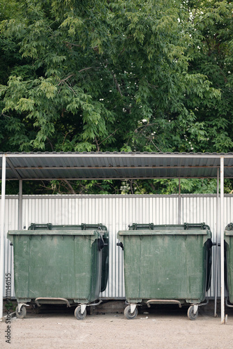 Outdoor waste containers under a canopy in a residential yard. Green plastic bins on wheels by a metal fence and trees, ideal for urban infrastructure and waste management projects.