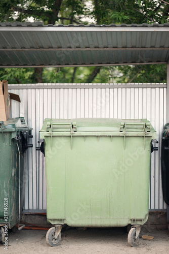 Outdoor waste containers under a canopy in a residential yard. Green plastic bins on wheels by a metal fence and trees, ideal for urban infrastructure and waste management projects.