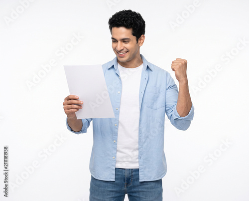 young attractive indian man celebrating while holding blank a4 paper isolated on white background for banking finance salary bonus tax refund and investment success concepts