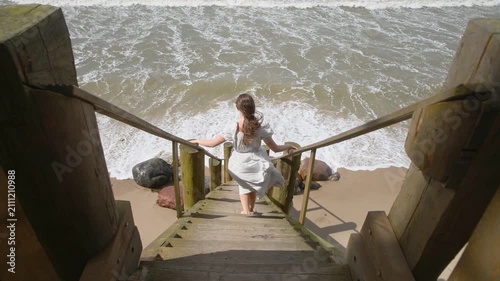 Woman in white dress descending wooden stairs to rocky seaside with crashing waves midday