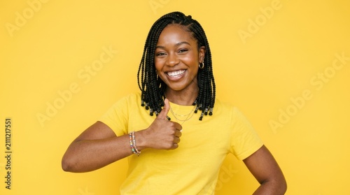 A smiling Black woman with braided hair is giving a thumbs-up gesture. She is wearing a yellow t-shirt and a beaded necklace.