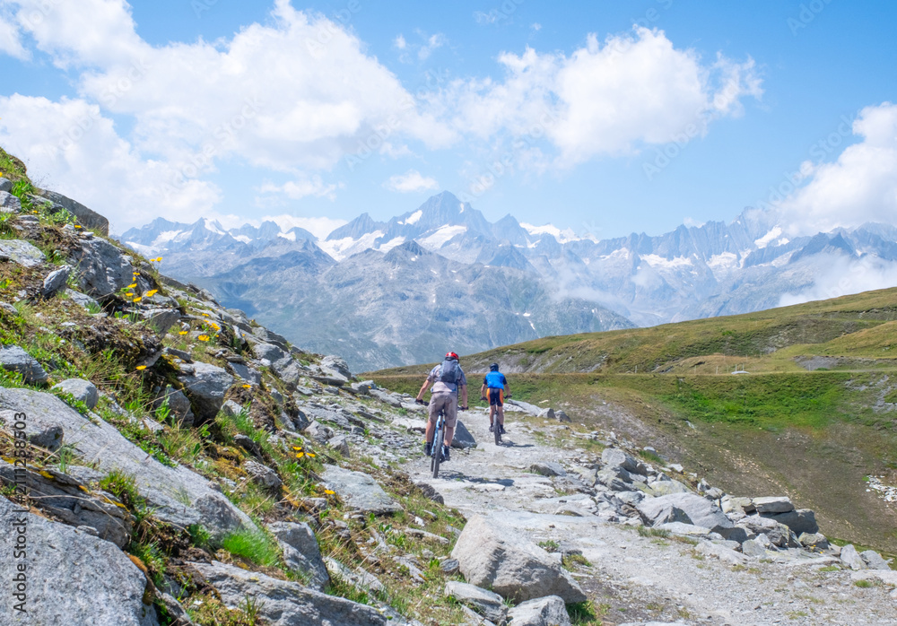 Fototapeta premium Rear view of two unrecognizable mountain bikers cycling along a rugged rocky trail with expansive alpine scenery and distant snow-covered peaks in Switzerland