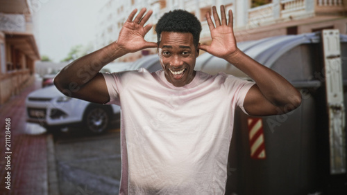 Foto Man raising hands by head and smiling, wearing pink tshirt beside dumpster on street; playful energy mischief