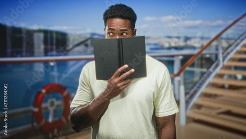 Canvastavla Man holds book over mouth and peeks with hand visible on building deck near lifebuoy and stairs; curiosity contemplation