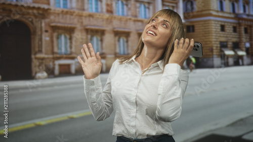 Fotografie Woman holding smartphone to ear, smiling with hand raised while standing on a city street; joyful connection