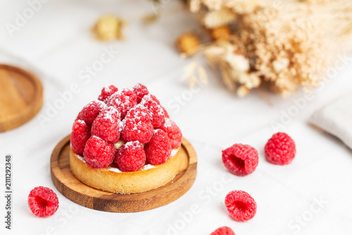 Konstfotografi Raspberry tartlet on a wooden plate lying on a beige napkin sprinkled with raspberries, alongside dry flowers