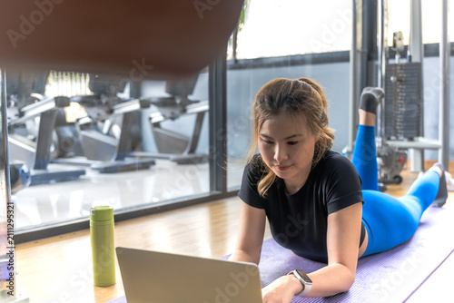 Foto Asian sportswoman lying on fitness center floor working on laptop looking for ex