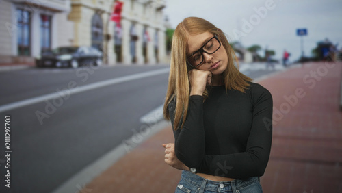 Foto Woman with glasses rests chin on hand on street near building, wearing black shirt and jeans in a reflective pose; quiet contemplation