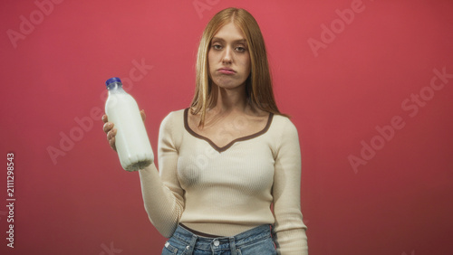 Принти на полотні Redhead woman holds a white milk bottle in one hand, puckering lips and rolling eyes while wearing a cream sweater and jeans in a studio red backdrop; skepticism taste testing