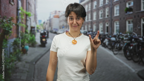 Canvas Print Woman gesturing with hands in white t-shirt and orange pendant on a narrow street lined with bicycles and brick rowhouses, squinting and grimacing; playful movement