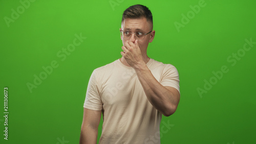 Quadro su tela Man pinching nose with right hand and gesturing with left hand while grimacing in a green studio wearing glasses and beige shirt; disgust