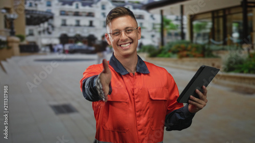 Cuadro en lienzo Man mechanic in orange coverall offering handshake with bare right hand while holding tablet in front of building entrance; friendly welcome
