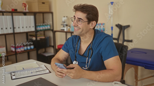 Man doctor holds smartphone and smiles in clinic building, stethoscope visible on his neck; calm reassurance.