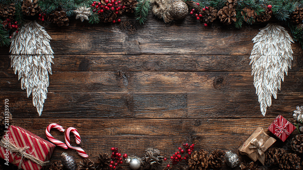 Obraz premium Festive Christmas background on dark wood featuring pine branches, cones, candy canes, red berries, angel wings, and gifts at top with copy space. 