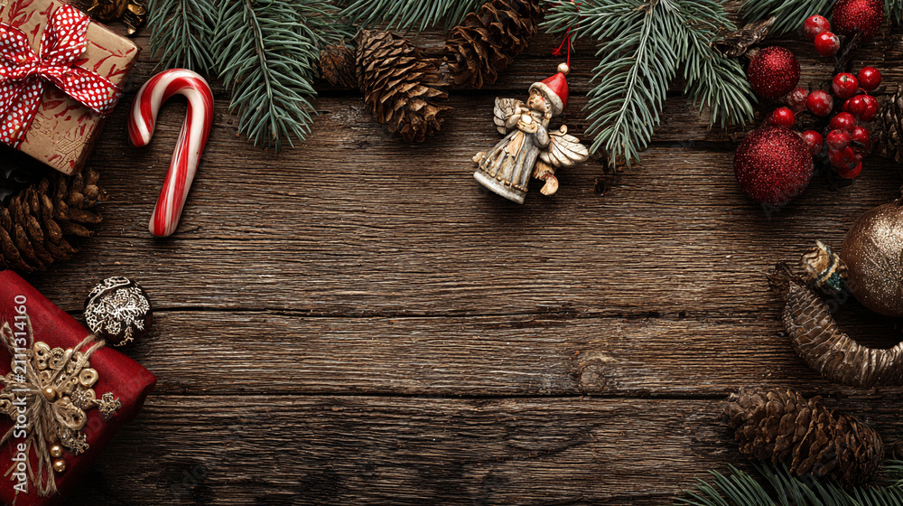 Obraz premium Festive Christmas background on dark wood featuring pine branches, cones, candy canes, red berries, angel wings, and gifts at top with copy space. 