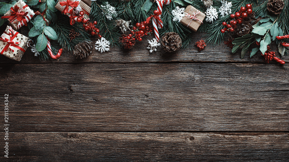 Obraz premium Festive Christmas background on dark wood featuring pine branches, cones, candy canes, red berries, angel wings, and gifts at top with copy space. 
