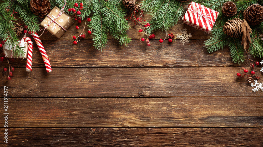 Obraz premium Festive Christmas background on dark wood featuring pine branches, cones, candy canes, red berries, angel wings, and gifts at top with copy space. 