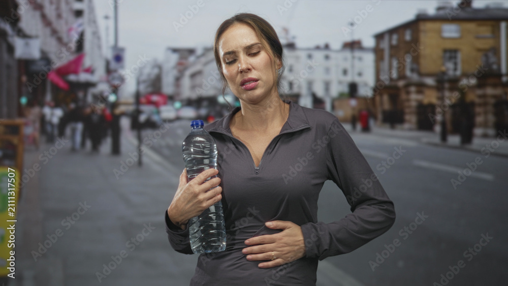 Fototapeta premium Woman hispanic holding a large water bottle, hand on stomach indicating stomachache on a busy urban street; discomfort nausea.