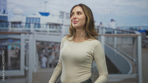 Canvas Print Woman gazes upward with a soft smile by ship railing on a building deck wearing a beige longsleeve top and jeans; serenity contemplation