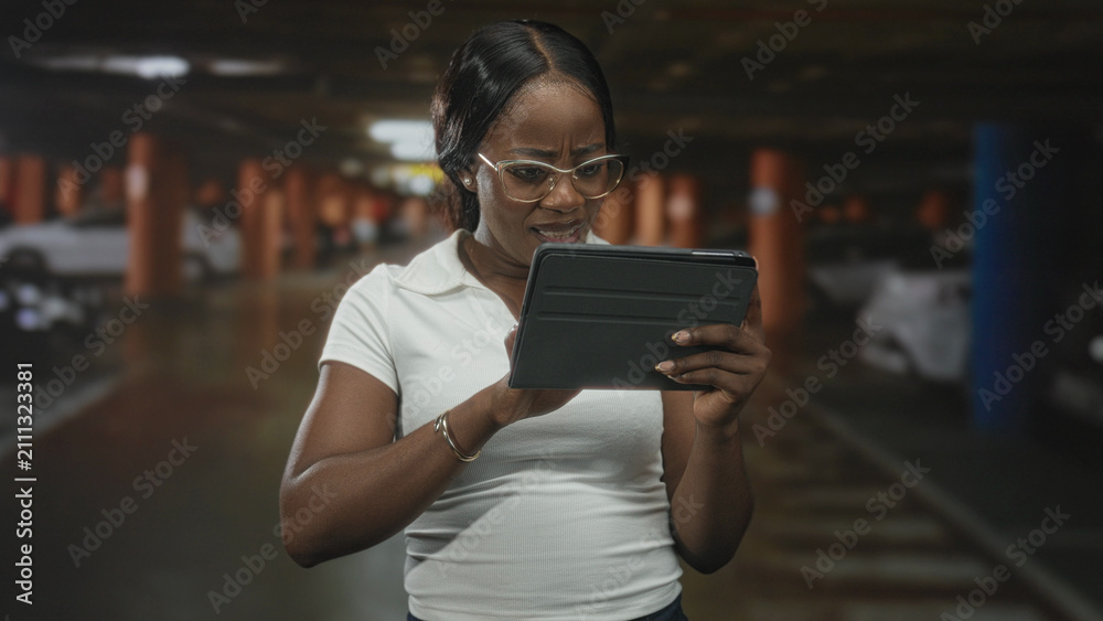 Fototapeta premium Woman holds tablet with both hands, tapping screen with right hand in a dim parking building, wearing glasses and white polo; concern focus.