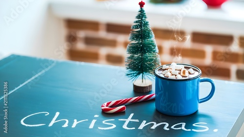 Christmas concept with a blue mug of hot chocolate with marshmallows, a small decorative pine tree, and a candy cane on a dark table with the word christmas written in chalk