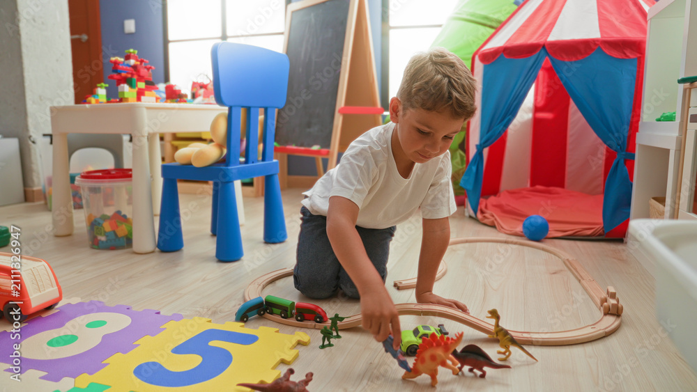 Fototapeta premium Boy kneeling and reaching with hand toward toy train on wooden floor in building playroom with tent, chair and foam puzzle mat; imaginative play joy.