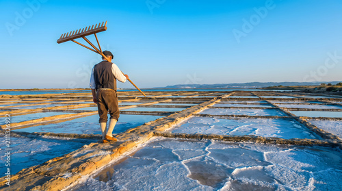 Salt field worker carrying a rake on shoulder walking across salt pans
