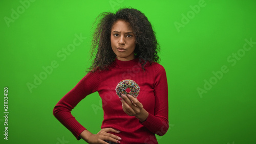 Konstfotografi Woman with curly hair holding chocolate doughnut with hand on hip in studio, red sweater, glancing aside; contemplation