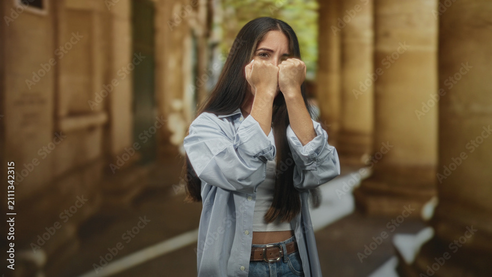 Fototapeta premium Woman with fists raised covering her face in a building colonnade walkway, hands tense and protective; anxiety resilience guarded determination.