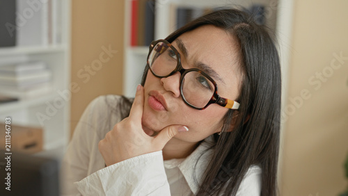 Fotografía Woman wearing tortoiseshell glasses with hand on chin and pensive expression in office with shelves, binders and storage boxes; contemplation