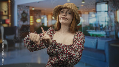 Foto Redhead woman pointing to wrist and tapping with fingers, frowning in a hotel lobby building; impatience dismissal