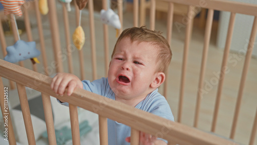 Foto Boy crying with mouth open, gripping wooden crib rail and reaching toward hanging mobile in nursery; distress separation