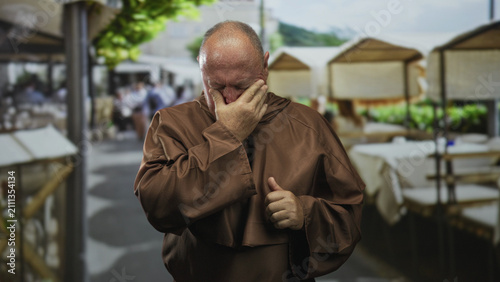 Foto Man monk in brown robe covers face with hand and wipes tears while standing by outdoor cafe stalls on street; sorrow reflection repentance