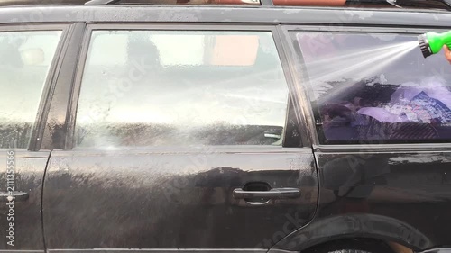 Person cleaning car window with hose spray, woman washing vehicle window with water nozzle outdoors, car maintenance.