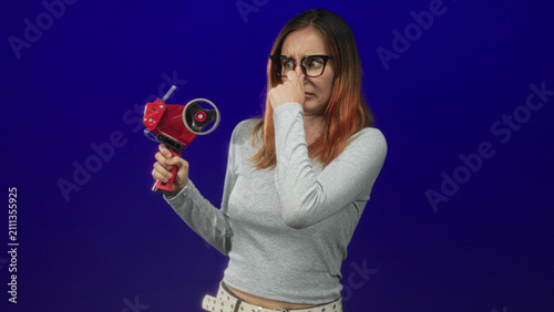 Obraz na płótnie Woman pinches nose with fingers while holding red tape dispenser and wearing glasses in studio under bright lights; disgust