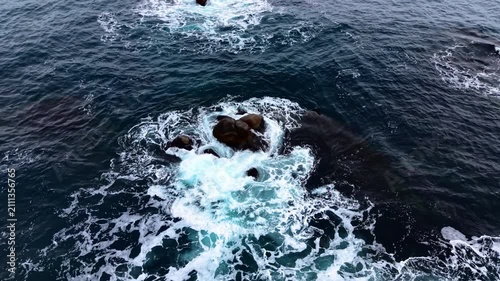 Overhead abstract view of waves swirling around rock. Drone shot of dark blue ocean, white foam and a single boulder creating natural pattern.