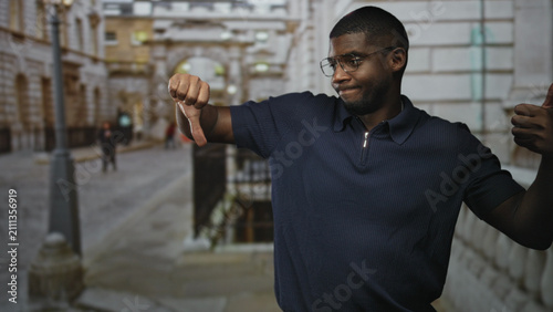 Принти на полотні Young black man in navy polo gives left thumb down and right thumb up on a stone street while wearing glasses and a slight smirk; ambivalence