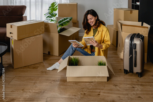 Картината върху платно Young woman unpacking boxes and smiling while settling into new home with suitca