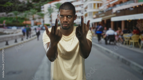Foto Man wearing beige tshirt shows both middle fingers with bare hands visible while facing camera on a street cafe terrace; defiance