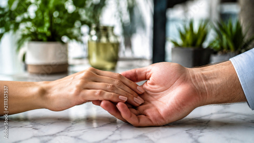 A Tender Connection: Two hands gently clasp in a moment of intimacy and support, surrounded by the soft ambiance of an indoor setting with lush greenery and dappled light.