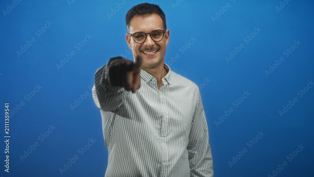 Naklejka premium Man with moustache in striped shirt smiles and points finger at camera, hand visible in studio backdrop with blue wall; playful charm.