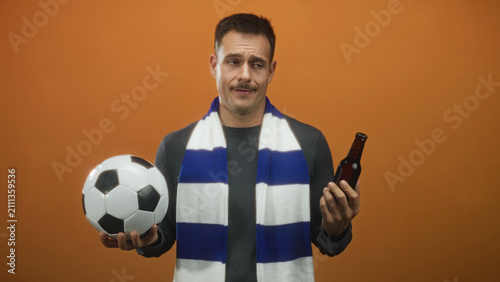 Man holding a soccer ball in one hand and a beer bottle in the other, hand in hesitant shrug, studio; uncertainty fandom dilemma.