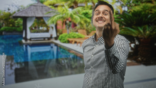 Фотография Young man with moustache points and shows middle finger toward camera by poolside building; defiance