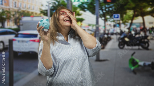 Foto Senior hispanic woman holds blue piggy bank with hand to face gesture on crowded city street; financial relief