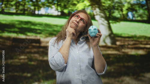 Konstfotografi Woman holds blue piggy bank and touches chin while considering savings, standing in a forest park by terraced stone steps; retirement worry