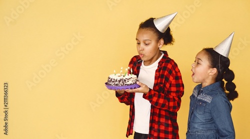 Two young diverse girls in party hats standing against a yellow background, one blowing out candles on a birthday cake while the other watches in excitement.