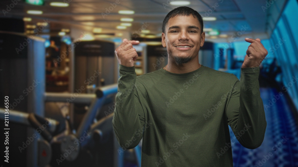 Fototapeta premium Man smiling and showing hands and face while making finger heart gesture at airport gate with terminal windows and seating; joy.